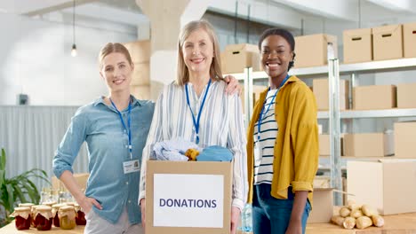 dos jóvenes y una voluntaria mayor sosteniendo una caja de donación con ropa y sonriendo a la cámara en un almacén de caridad