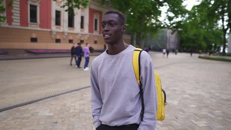 Positive,-Smiling-Young-Man-Walking-In-City-With-Yellow-Backpack-On-Shoulders,-Wearing-Casual-Clothes