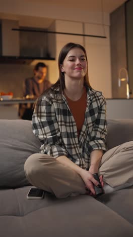 vertical video of a happy brunette girl in a plaid shirt sitting on a gray sofa and switching channels on the tv using the remote control in a modern apartment at home near her boyfriend in the background who is working on his laptop