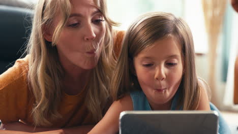 family with mother and daughter lying on floor at home pulling faces for selfie on digital tablet