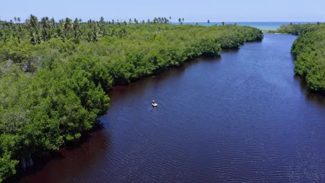 Vista-Aérea-De-Un-Pequeño-Bote-Navegando-En-Un-Río-Idílico-Rodeado-De-Bosques-De-Manglares-Durante-El-Verano---Mar-Caribe-Tropical-En-Segundo-Plano