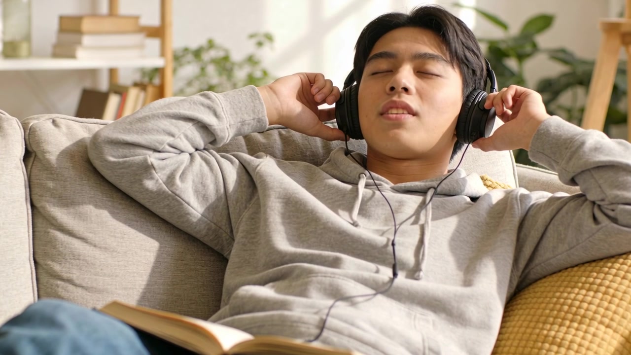 ライトグレーのパーカーを着た男性がソファに横たわり、黒い有線ヘッドホンを持ち、勉強しながら聞いている。