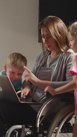 curious children point fingers to screen of laptop distracting freelance mom from work. woman with spinal cord injury sits in wheelchair near kids