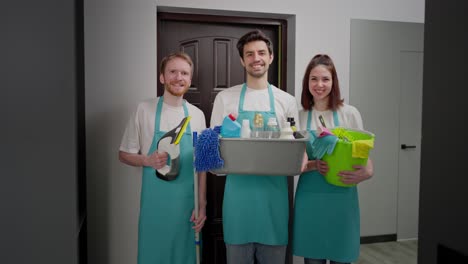 retrato de un feliz trío de limpiadores en frentes azules con varias herramientas y un limpiador para limpiar en sus manos dos hombres y una chica morena en un apartamento moderno en una llamada de limpieza