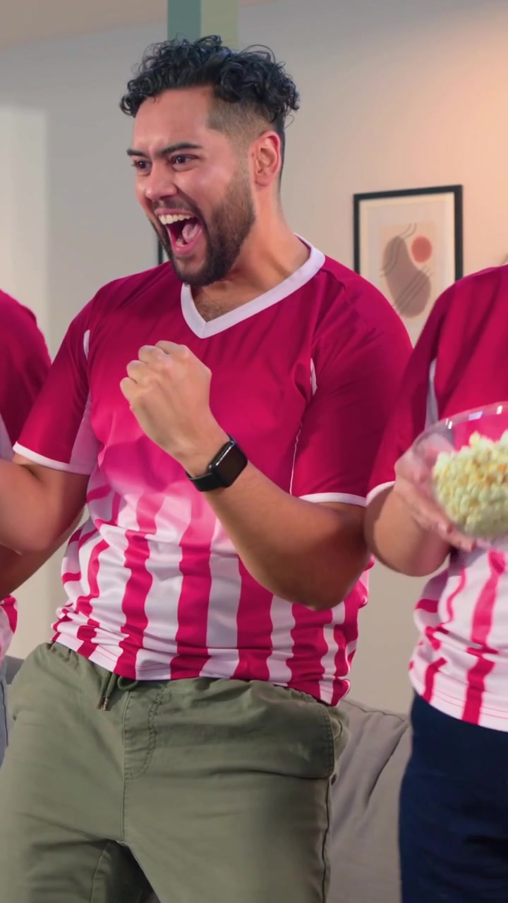 Video vertical: Después de un gol en la televisión, hombre con camiseta a rayas moviendo el balón, agarrando palomitas de maíz y animando en casa.