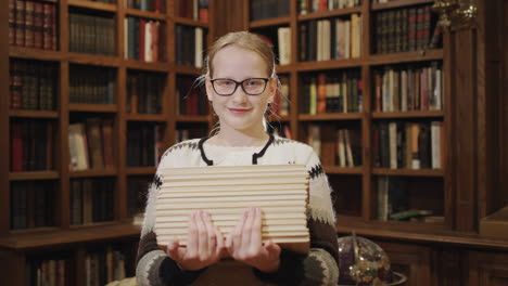 Portrait-of-a-schoolgirl-with-a-stack-of-textbooks-in-her-hands,-stands-against-the-background-of-racks-with-books-in-the-library.