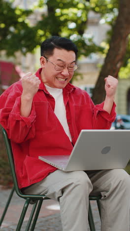 Asian-happy-man-working-on-laptop-celebrate-success-win-money-sitting-on-urban-street-in-city