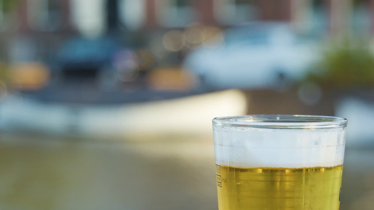 Hand lifts clear pint of golden beer outdoors by river, with blurred background in daylight