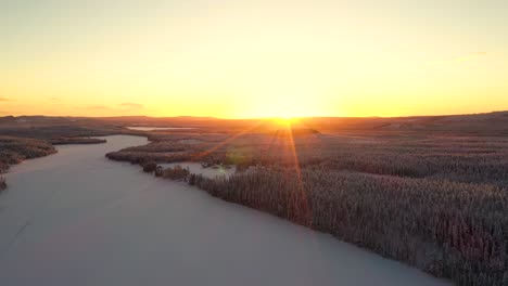 Vista-Aérea-De-La-Puesta-De-Sol-Sobre-Un-Lago-Nevado-Congelado-En-Un-Bosque-Profundo-Con-Colinas-Y-Montañas