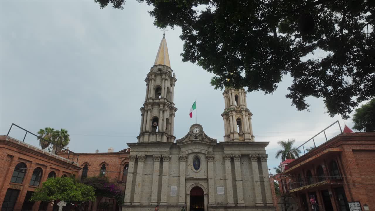Establishing view of Iglesia San Francisco de Asis, Chapala’s iconic church spire framed by lush trees under a cloudy sky in Jalisco, Mexico