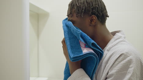 immigrant man in bathrobe covers his face with blue towel in modern bathroom. close-up of his hands holding towel, showing self-care or freshening up. minimalistic bathroom setting