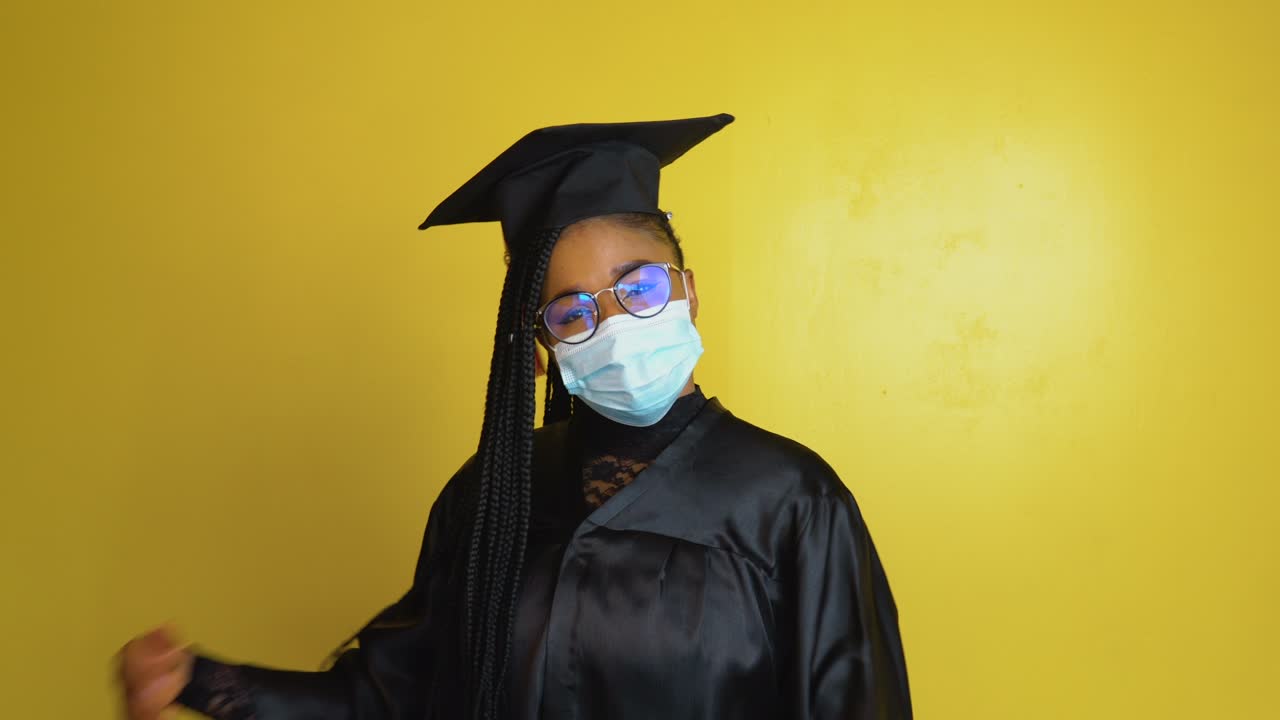The graduate student in a protective mask expresses with gestures the need to comply with quarantine security measures. Student in a black robe and hat on a yellow solid background looks at the camera
