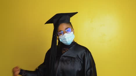 el estudiante graduado con una máscara de protección expresa con gestos la necesidad de cumplir con las medidas de seguridad de cuarentena. estudiante con una túnica negra y sombrero sobre un fondo sólido amarillo mira a la cámara