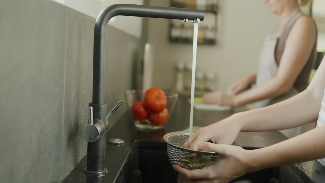 Mamá e hija preparan una ensalada en la cocina. En primer plano, unas manos lavan una ensalada; al fondo, una mujer las corta en una tabla de cortar.
