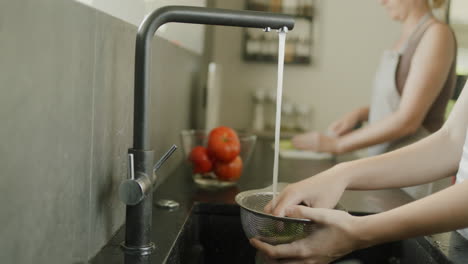 Mom-and-daughter-are-preparing-a-salad-in-the-kitchen.-In-the-foreground,-hands-are-washing-a-salad,-in-the-back,-a-woman-cuts-them-on-a-cutting-board