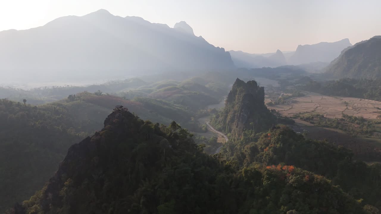 Imponenti creste calcaree dominano il paesaggio di Nam Xay a Vang Vieng, Laos, circondate da vaste terre agricole, strade tortuose e montagne stratificate in lontananza sotto una luce atmosferica morbida e dorata