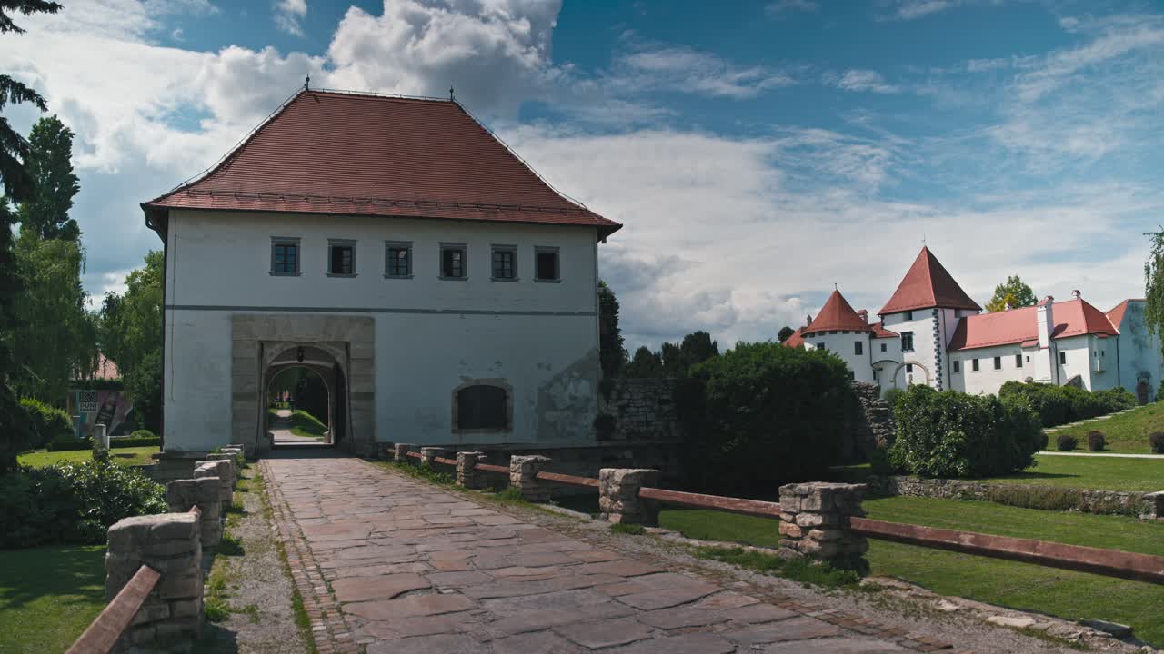 Storico castello di Varazdin e un sentiero lastricato in pietra che conduce attraverso un cancello ad arco, circondato da una lussureggiante vegetazione e un cielo azzurro brillante, Croazia.