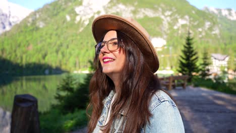 Mujer-Con-Sombrero-Marrón-Gira-Y-Sonríe-A-La-Cámara-En-La-Zona-Boscosa-De-Lago-Di-Braies,-Italia