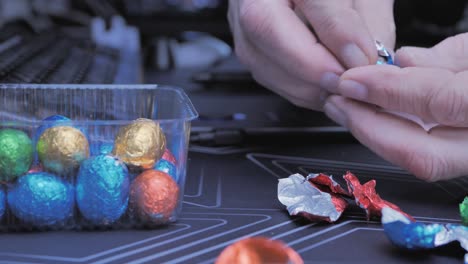 adult man eating chocolate easter eggs while working on computer at home, focus on hands taking colorful snacks