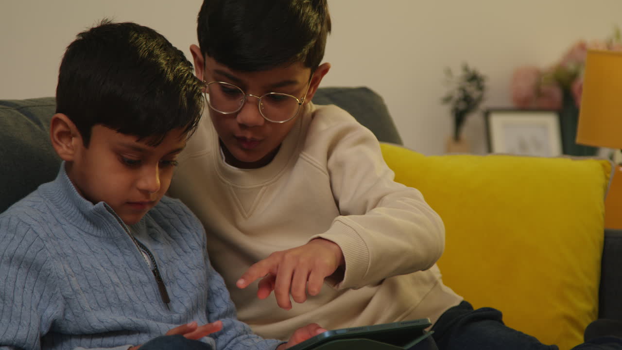 Two Young Boys Sitting On Sofa At Home Playing Games Or Streaming Onto Digital Tablet Together 1