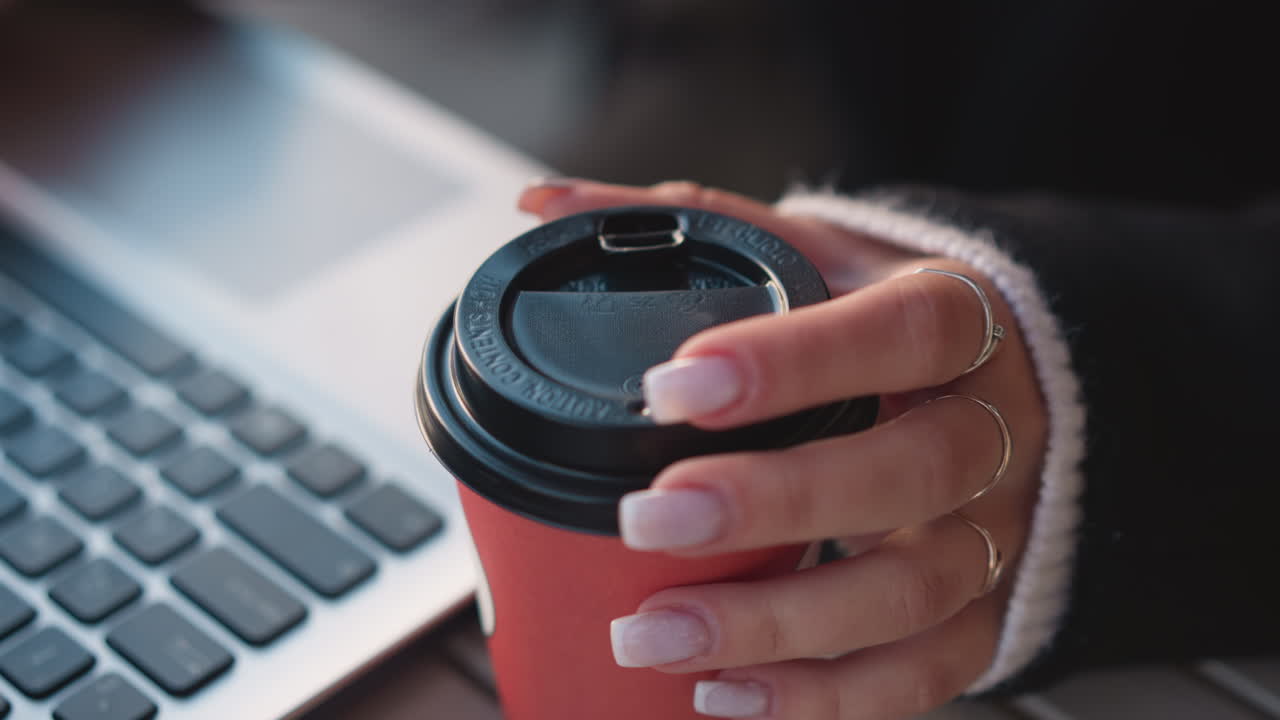 여성의 손이 노트북 근처에 커피 컵을 부드럽게 돌리는 클로즈업으로, 흐릿한 키보드 배경에 매니큐어 된 손가락에 초점을 맞추고 있습니다.