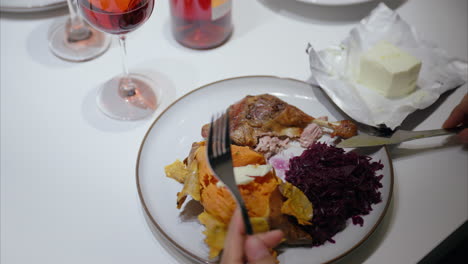 woman has healthy dinner at home, using fork and knife, cutting meat, roasted duck leg, sweet potato, red cabbage, balanced diet, festive, delicious meal, homemade dish, enjoying food with red wine