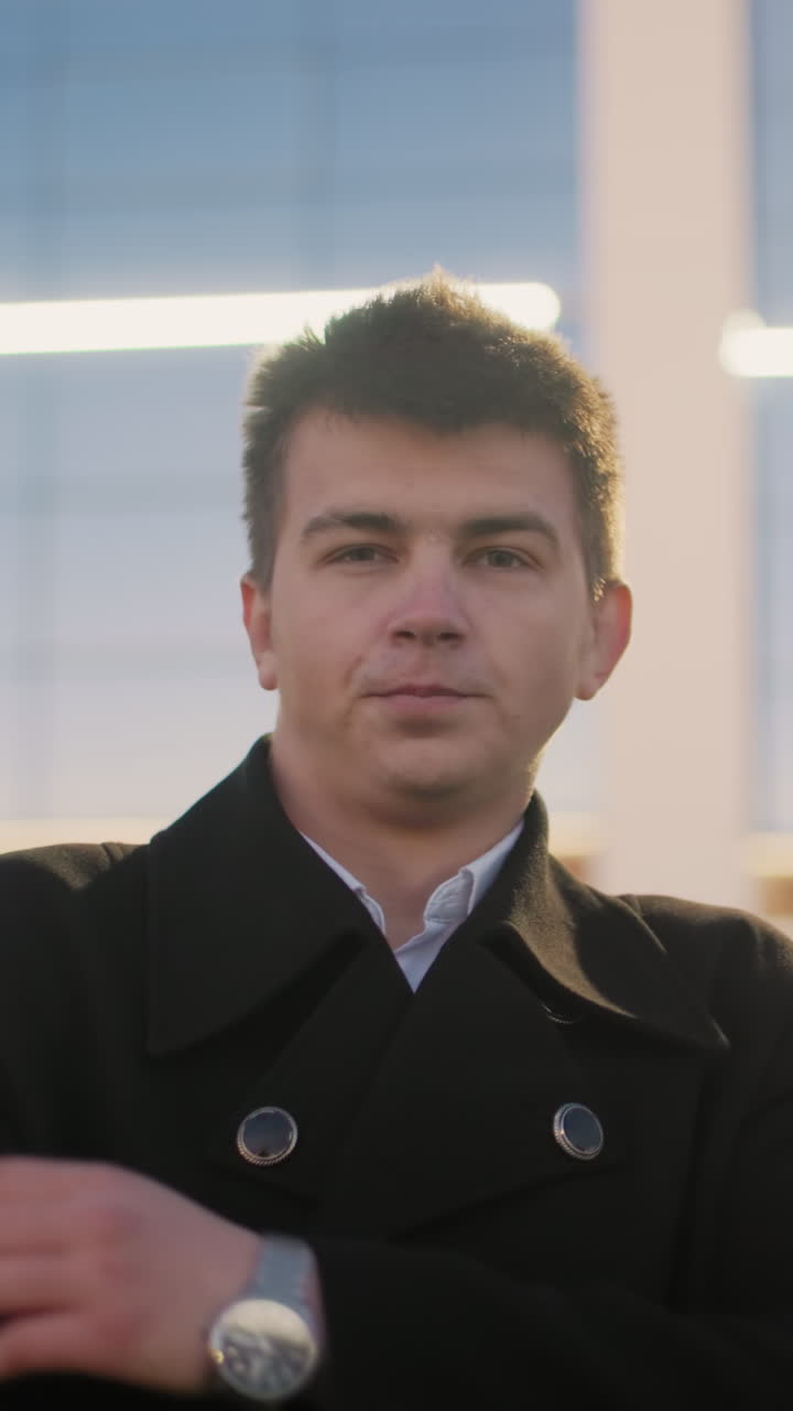 человек, стоящий на открытом воздухе перед торговым центром, случайно касаясь подбородка, с стеклом торгового центра, отражающим золотой свет и структуру, создавая расслабленную, вдумчивую атмосферу в городской обстановке