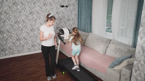 indoor workout session featuring young girl running on treadmill as female coach monitors her progress with outdoor scenery faintly visible through large window beside sofa in modern living room