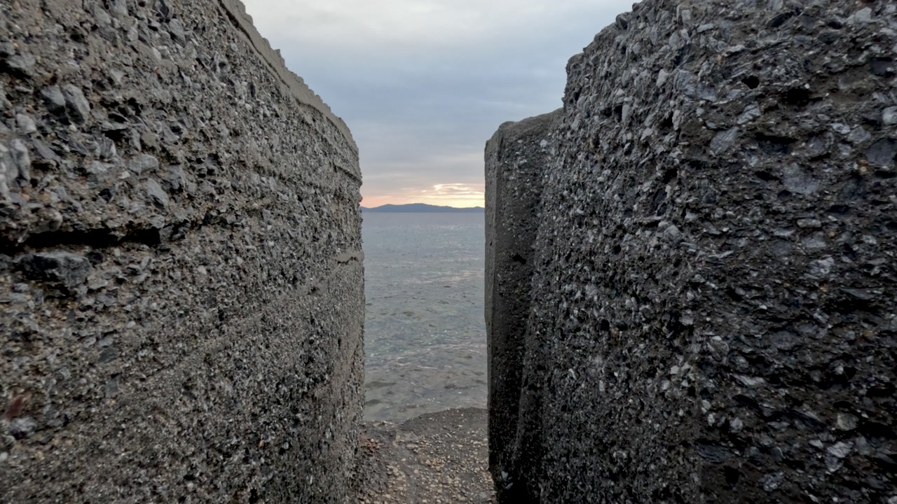 Two large concrete structures frame a view of the water and the sunset. The gap reveals a wide ocean view with distant land in the background, showcasing natural scenery.