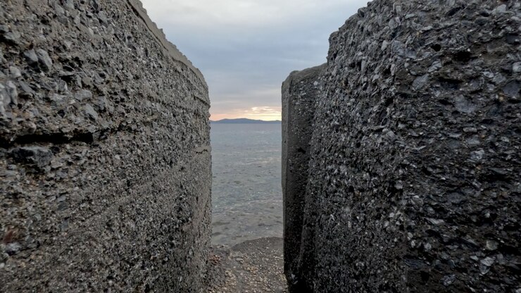Two large concrete structures frame a view of the water and the sunset. The gap reveals a wide ocean view with distant land in the background, showcasing natural scenery.