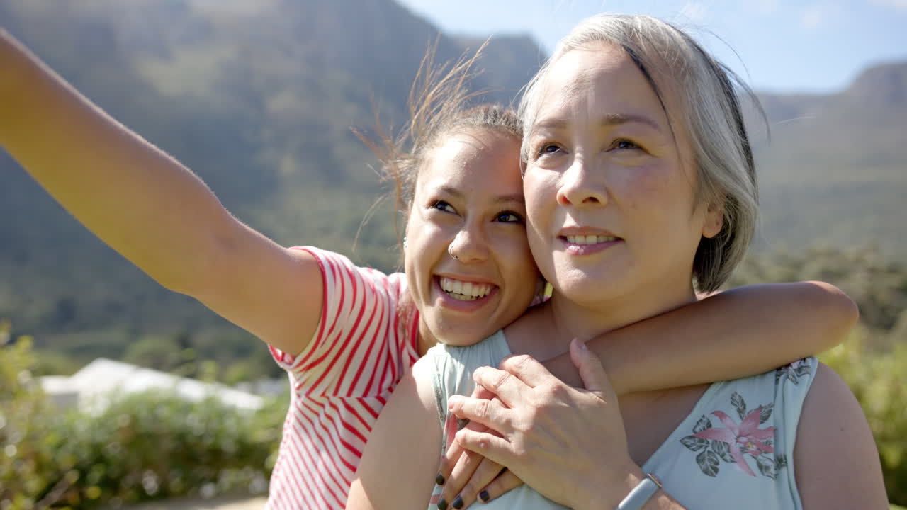 Smiling young asian woman hugging asian grandmother and taking selfie outdoors in nature