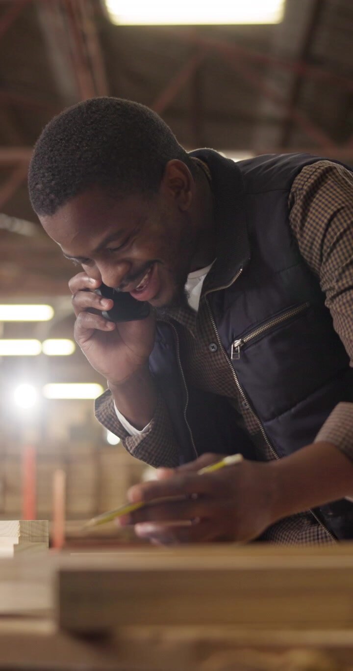 Carpenter Talking on Phone While Measuring Wood in Workshop