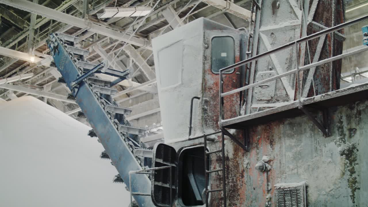 A tilt-up shot reveals a massive, old industrial reclaimer machine inside a factory warehouse, surrounded by huge piles of white granular fertilizer or chemical powder