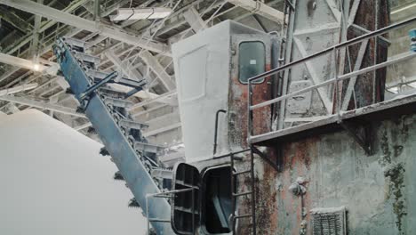 a tilt-up shot reveals a massive, old industrial reclaimer machine inside a factory warehouse, surrounded by huge piles of white granular fertilizer or chemical powder
