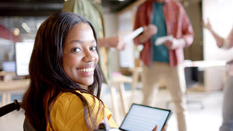 Happy-african-american-female-creative-in-wheelchair-using-tablet-in-casual-office-meeting