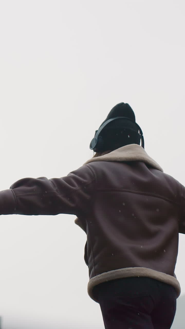 Back shot of smiling lady wearing black knit cap, brown shearling jacket, black trousers, arms outstretched while gracefully walking on wide stone path outdoors, enjoying cool autumn day