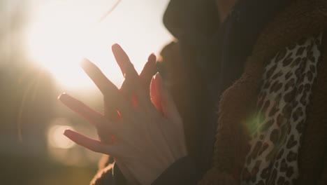 close-up shot of a person rubbing their hands together near their chest, lost in thought. the face is not visible, but the person is wearing a brown coat and hoodie. sunlight in the background