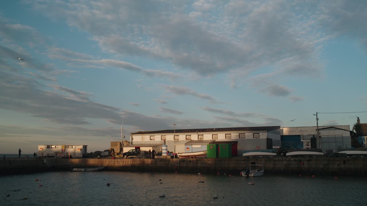 A locked-off shot of Bullock Harbour in Dublin, Ireland, during sunset. Fishing boats, a stone pier, and coastal buildings are illuminated by warm golden light under a soft blue sky.