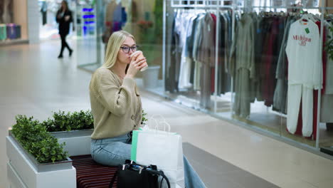 lady sipping coffee thoughtfully while seated near decorative flowers, background shows a clothing store with shelves filled with garments and a shopper walking by