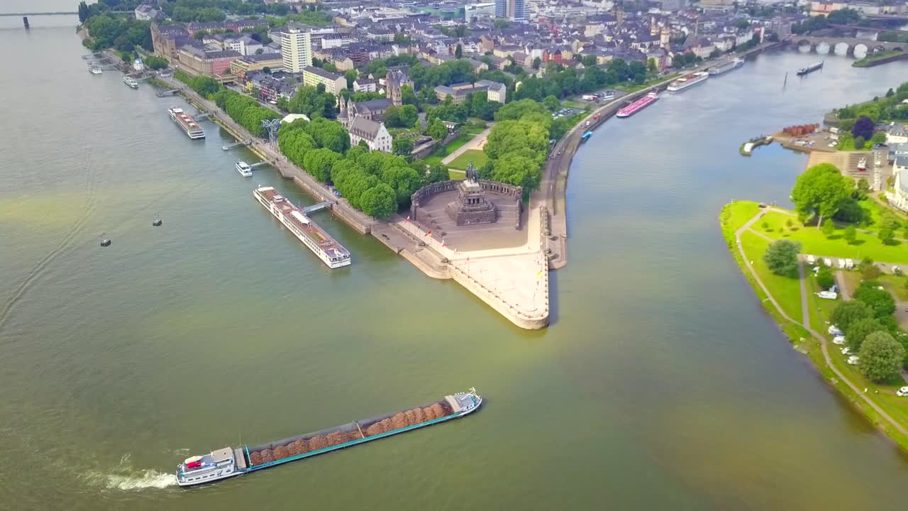 Stunning drone footage of Deutsches Eck in Koblenz, Germany, showcasing the confluence of the Rhine and Moselle rivers. Cargo boats navigate the waterways, adding movement to this iconic landmark