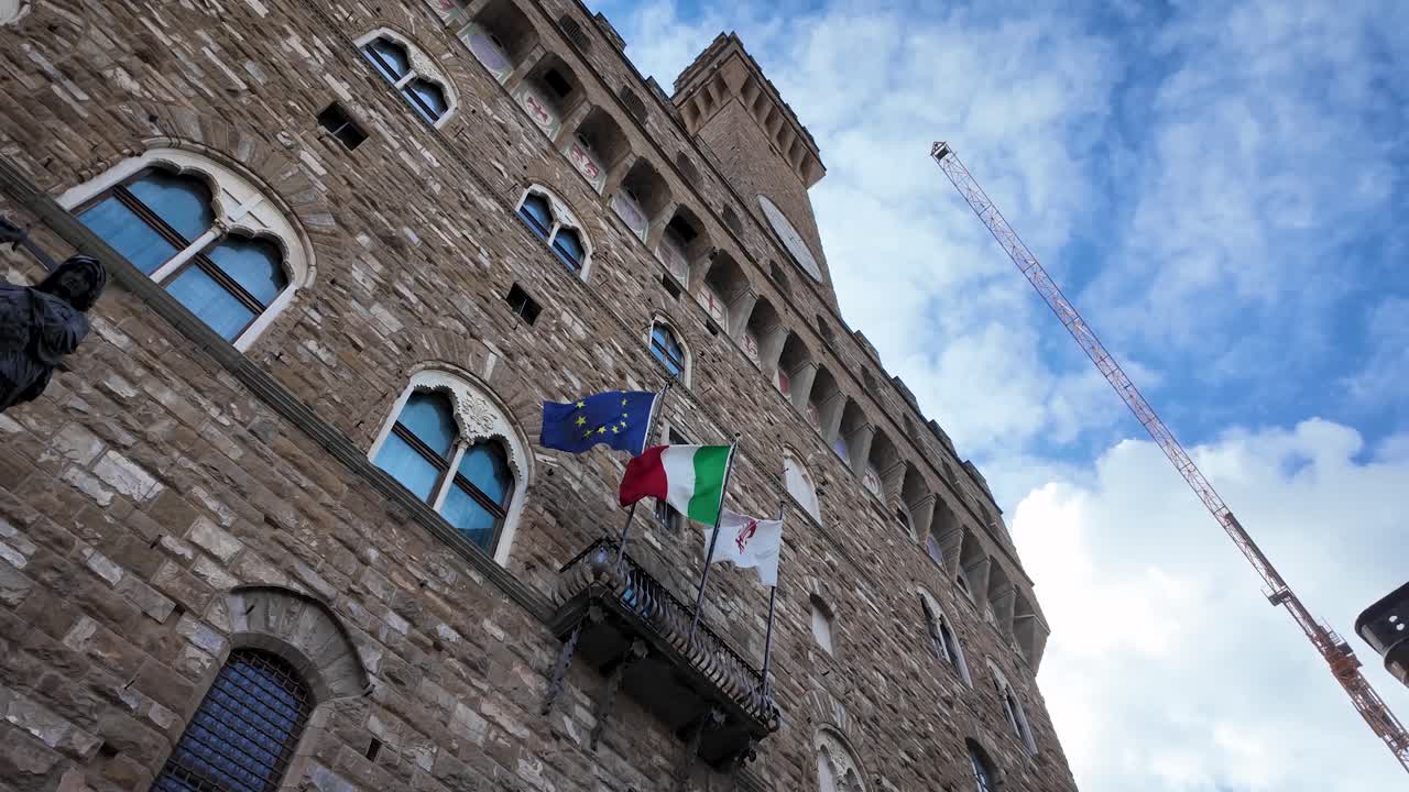 Vista zenitale statica della facciata principale di Palazzo Vecchio in una giornata di sole, che mostra le bandiere di Italia, dell'Unione Europea e di Firenze nel centro storico di Firenze, Italia.
