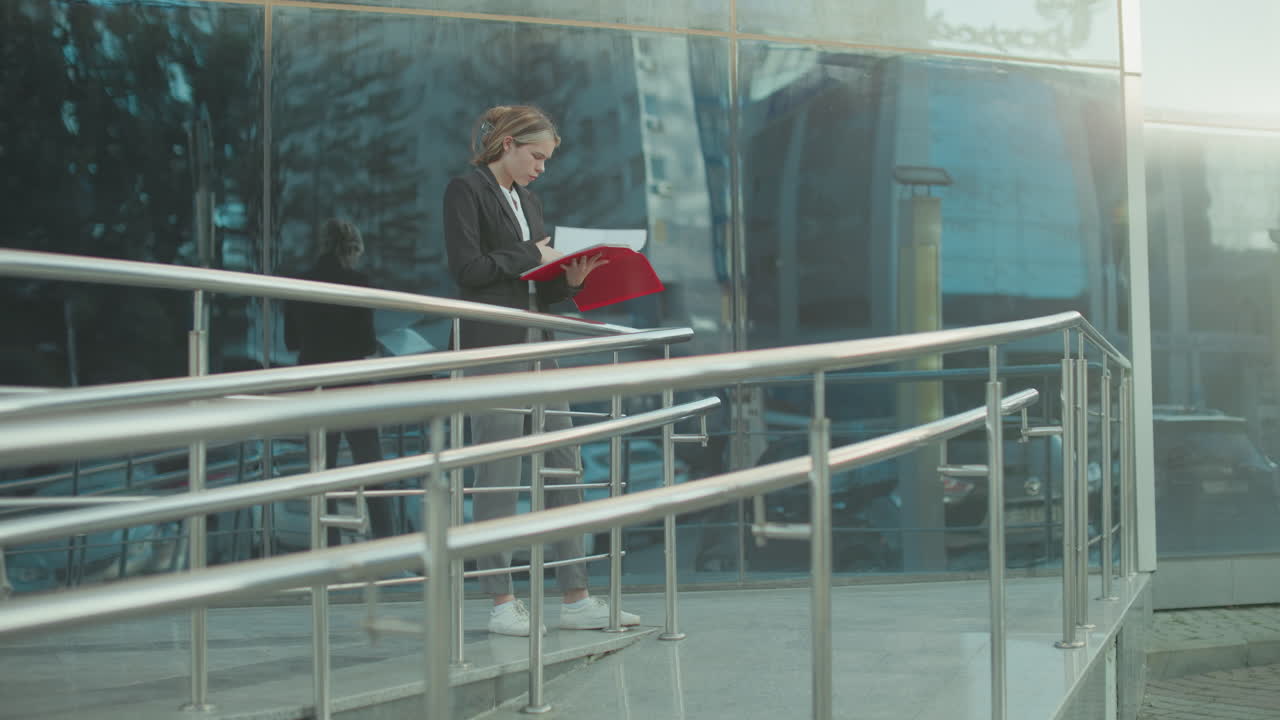 Young ceo reviewing red folder while standing beside iron railing outside glass office building reflecting urban surroundings with cars and structures
