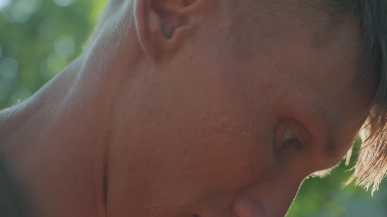 Closeup Profile Of Young Man Looking Down At Hands, Skin Texture And Jawline Visible, Soft Backlight From Trees, Intimate Shallow Depth Of Field Conveying Concentration And Momentary Absorption