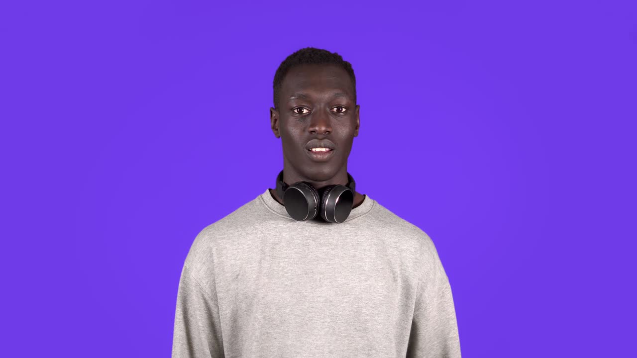 Cheerful young african man, gesturing in respect to someone. Isolated on blue background. Wearing white sweater and headphones on neck