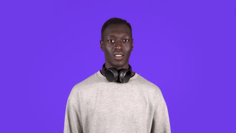 Cheerful-young-african-man,-gesturing-in-respect-to-someone.-Isolated-on-blue-background.-Wearing-white-sweater-and-headphones-on-neck