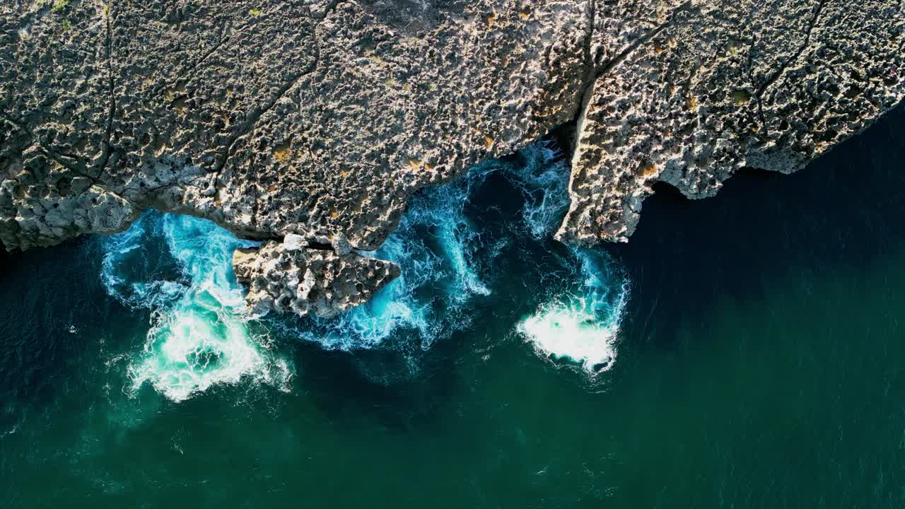 Cinematic overhead drone capture of a tropical rock coast where swirling waves meet sharp formations and golden sun reflection glows across shifting water in a raw untouched coastal environment
