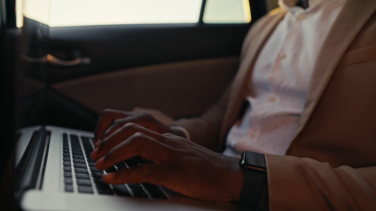 Close-up shot of a man Businessman with Black skin color and in a brown jacket works and types on a laptop while driving in the passenger seat in a modern car interior