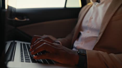 Close-up-shot-of-a-man-Businessman-with-Black-skin-color-and-in-a-brown-jacket-works-and-types-on-a-laptop-while-driving-in-the-passenger-seat-in-a-modern-car-interior