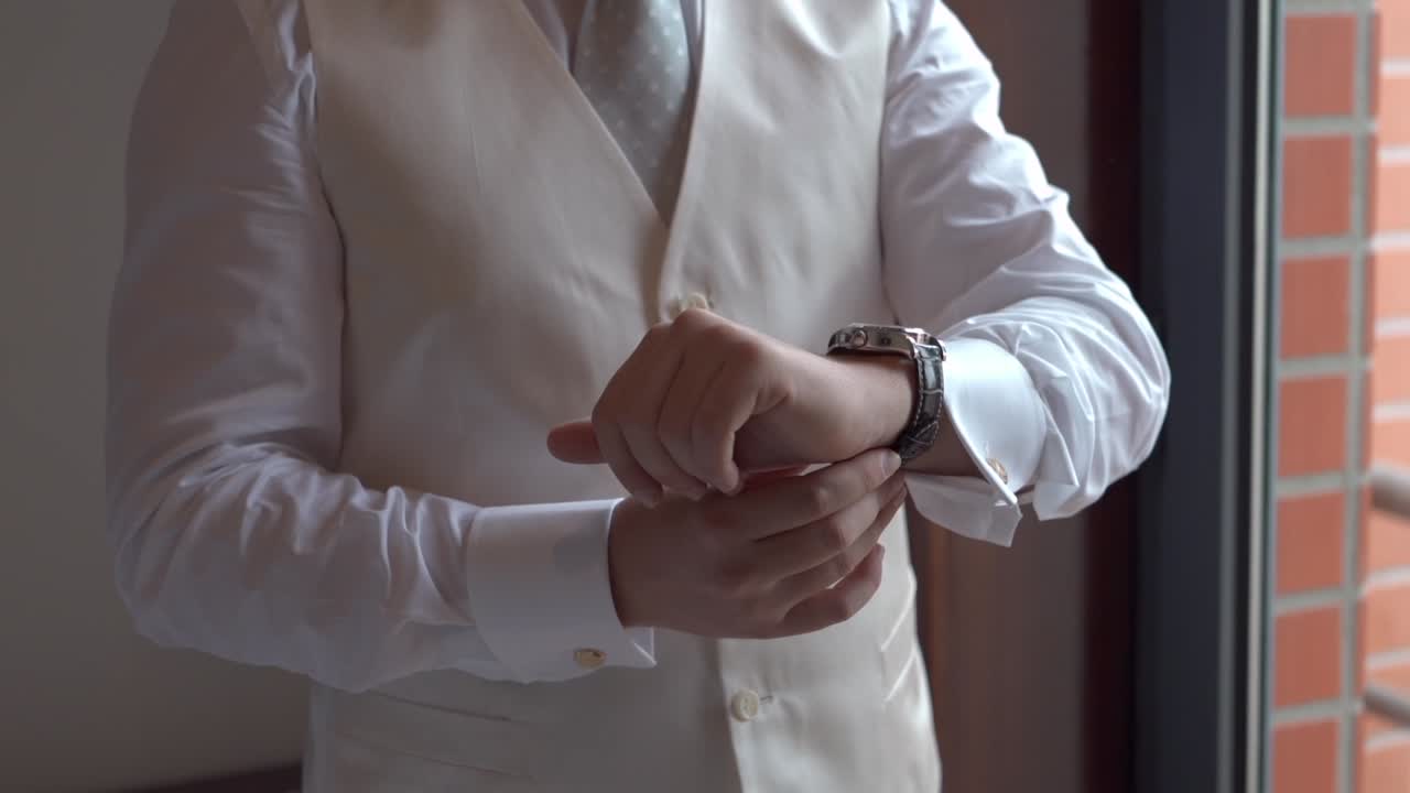 Groom in ivory vest and white shirt adjusting gold cufflinks and watch before wedding
