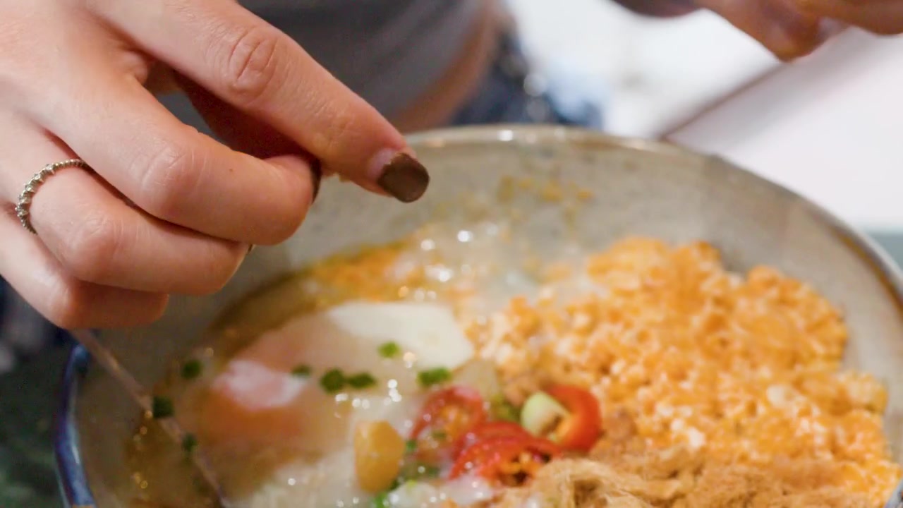 Primo piano di mani che spezzano pasta fritta in una ciotola di congee con uovo